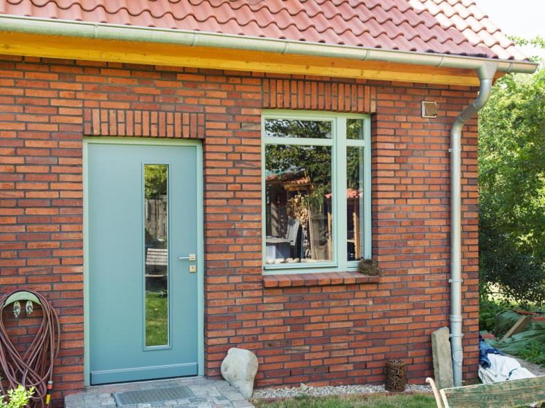 Türkise Holzhaustür mit länglichem Glasausschnitt neben türkisem Holzfenster